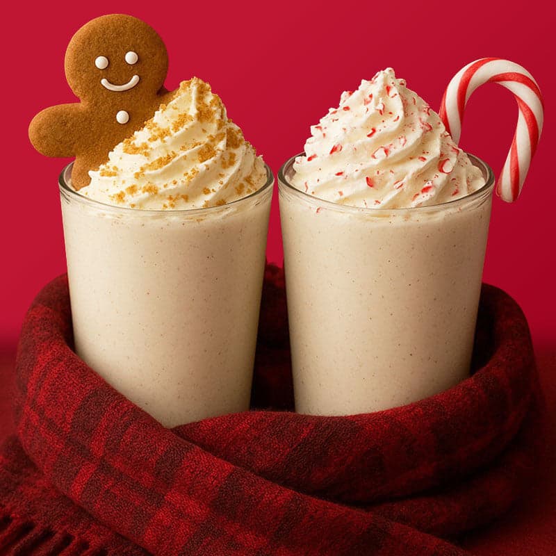 Two glasses of drinks with whipped cream. Gingerbread man and sprinkles on one, candy cane and flecks on the other. Wrapped in a red plaid scarf against a red background.