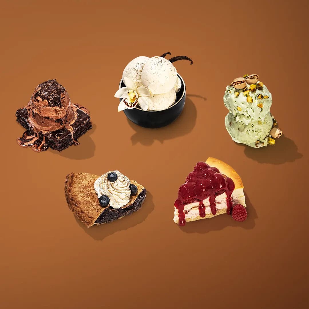 Five desserts on a brown background: chocolate brownie with melting ice cream, vanilla bean ice cream in a black bowl, pistachio ice cream, a berry pie slice with whipped cream, and a cherry-topped cheesecake slice.