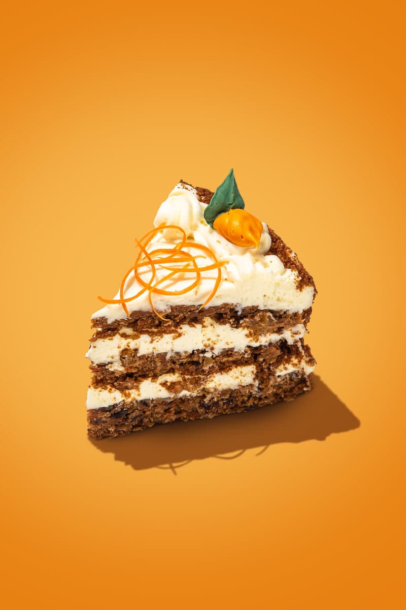 Slice of layered carrot cake with white cream cheese frosting, orange zest curls, and a small carrot and green leaf decoration, on an orange background.